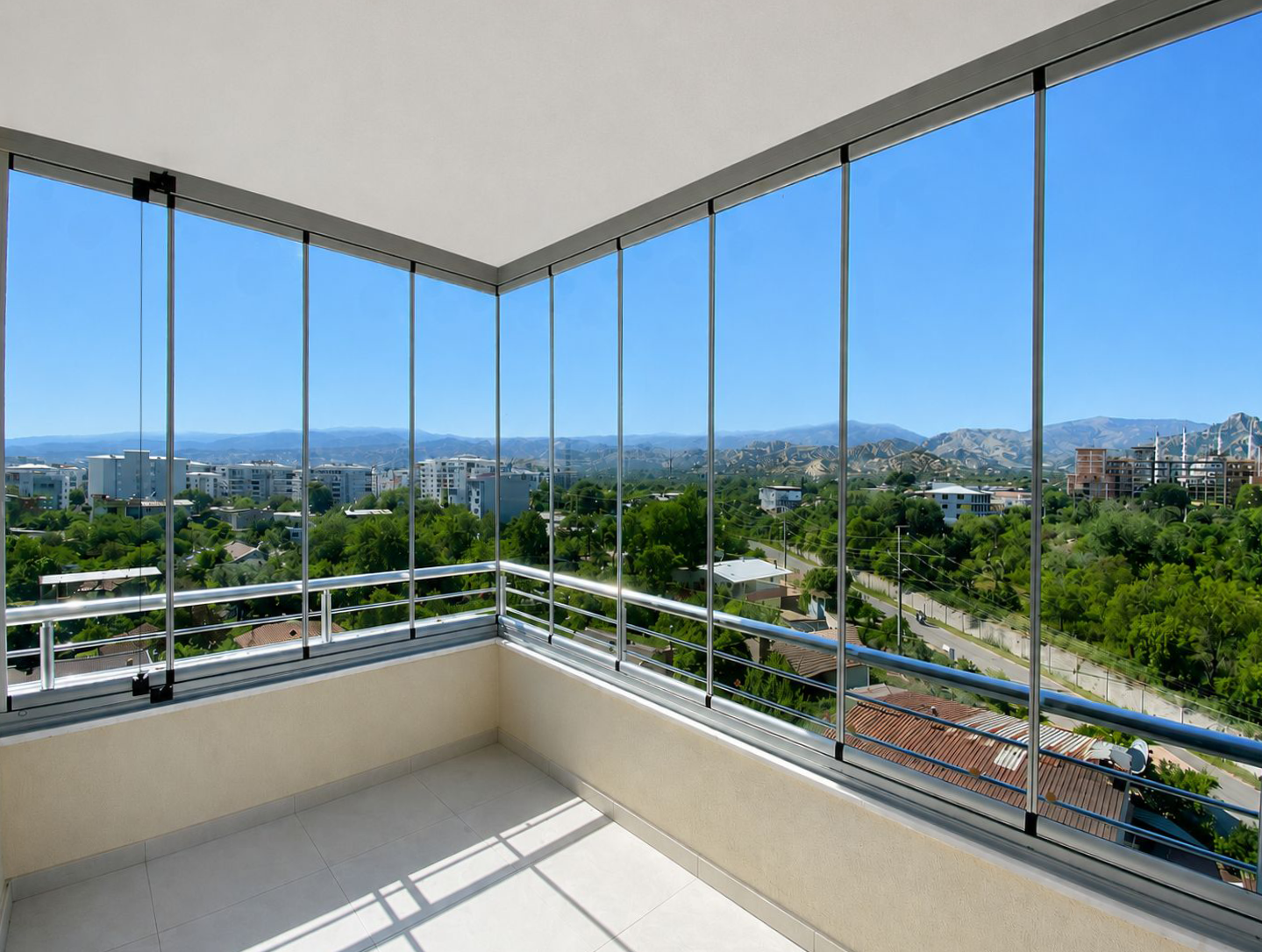 gold-plus-katlanır-cam-balkon-real-cam-balkon-05 Balcony with a glass railing overlooking a cityscape and distant mountains on a sunny day