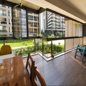 gold-plus-katlanır-cam-balkon-real-cam-balkon-07 Enclosed balcony with glass walls overlooking a green lawn and modern apartment buildings, wooden floor, and dining table with chairs.