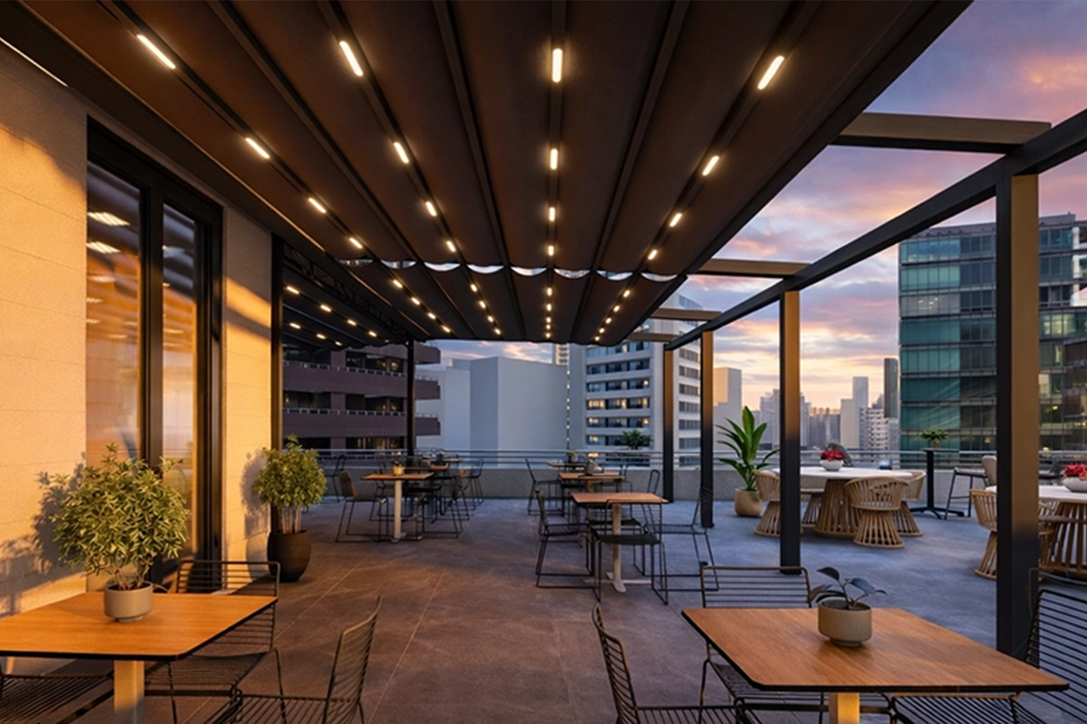 realcam-balkon-anasayfa-pergola Rooftop terrace with wooden tables and metal chairs, string lights, potted plants, and city skyline at sunset.